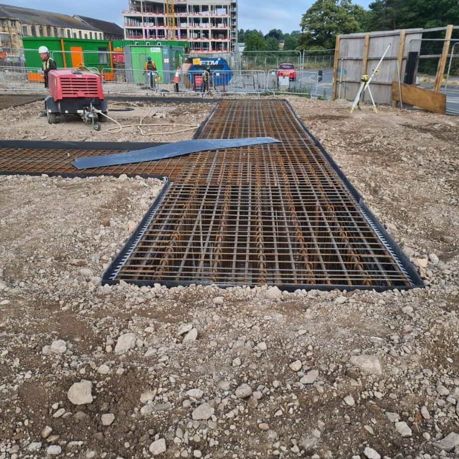 A construction site with rebar grids laid down on a gravel-covered ground, preparing for concrete pouring. A red machinery unit and several workers in safety gear are in the background near a partially constructed building and green construction containers.