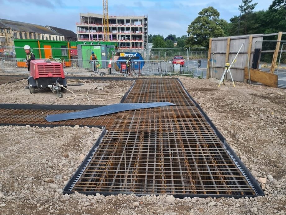 This image shows a construction site featuring a foundation grid of exposed rebar. Workers and various construction equipment, including a red generator, are visible. In the background, there are partially constructed buildings, temporary structures, and fencing.