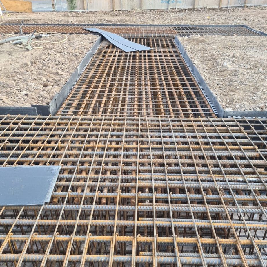 A construction site with a grid of rebar laid out, preparing for concrete pouring. Some rectangular sheets of metal are placed on top. The ground around the rebar is uneven with visible dirt and stones. Construction materials and tools are seen in the background.