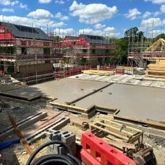 A construction site with two partially built houses surrounded by scaffolding. The foreground shows a freshly poured concrete foundation and various construction materials, including wooden planks and metal beams. The background features trees and a partly cloudy sky.