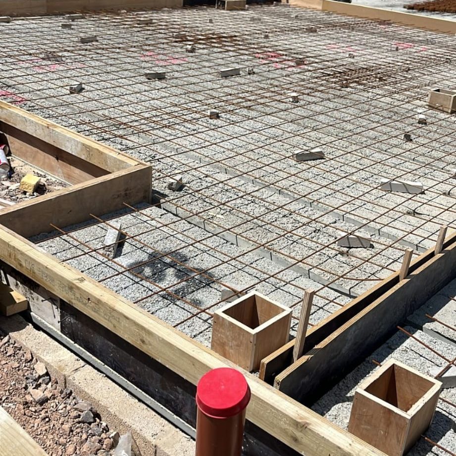 A construction site with a concrete foundation in progress. The area is framed with wooden beams and filled with a grid of rebar. Several small wooden boxes protruding from the concrete indicate future structural elements. Some tools and debris are scattered around.