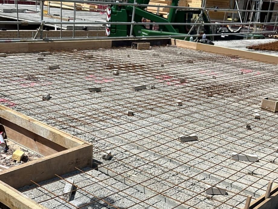 A construction site showing a concrete foundation being prepared with a grid of reinforcing steel rebar. The rebar is arranged in a crisscross pattern within a wooden frame. Various construction materials and equipment are visible in the background.