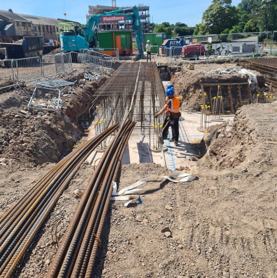 Construction site with workers building a structure. One worker wearing a blue helmet and an orange safety vest is standing near metal rebar and concrete forms. Machinery and materials are scattered around. Trees and buildings are visible in the background.