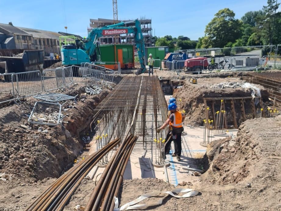 Construction site with workers in safety vests and hard hats preparing rebar framework for a foundation. A large green machine with "Plantforce" branding is on the site. Various construction materials and equipment are scattered around. Buildings and trees are in the background.