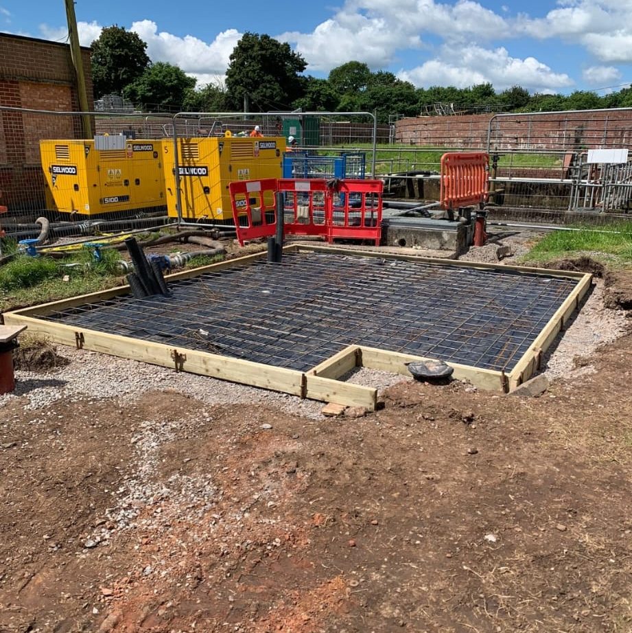 Construction site with a rectangular concrete foundation in progress. The foundation is framed with wooden boards and has reinforcing steel bars installed. Yellow and red construction barriers and equipment are visible in the background. Trees and a blue sky are behind.