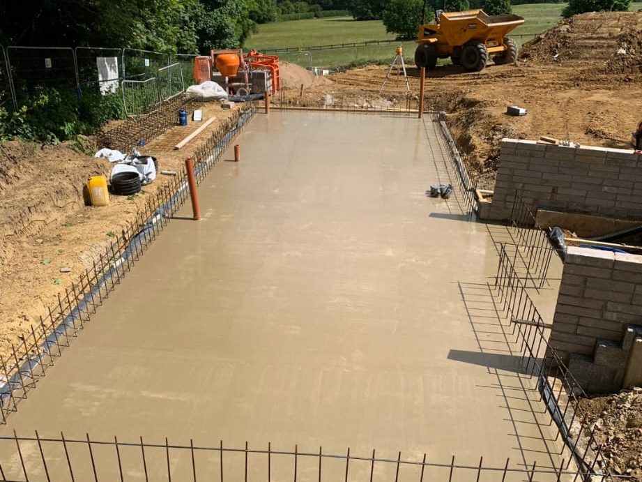 A construction site showing freshly poured concrete slab with rebar protruding from the edges. A small brick wall is being constructed on the right side, and construction equipment including a yellow dumper and a cement mixer is visible in the background.