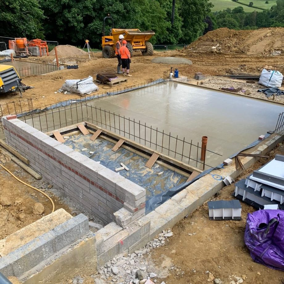 Construction site with a concrete foundation slab partially completed. Two workers are near a yellow dump truck in the background. Brick walls outline the foundation area, and construction materials are scattered around. Trees and hills are visible in the distance.