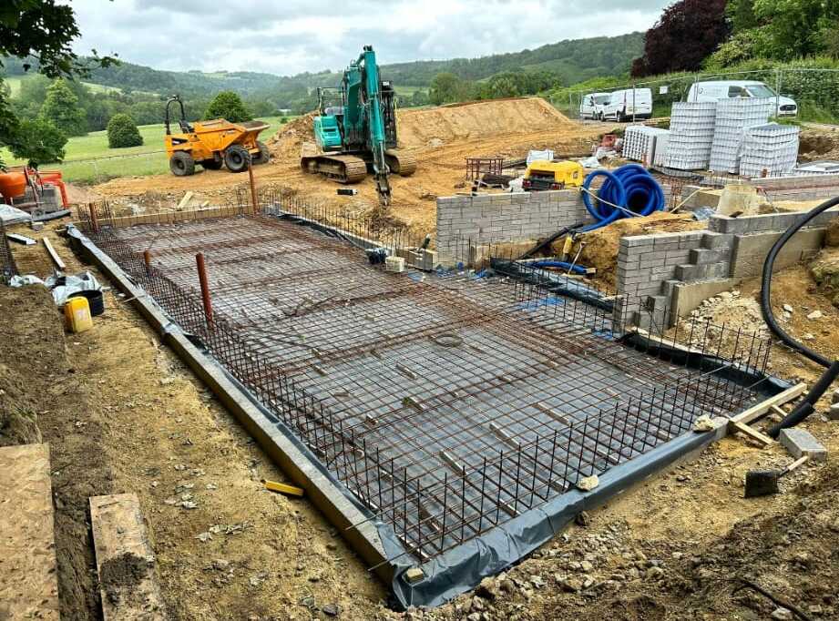 A construction site with a concrete slab foundation in progress. Metal rebar grids are laid out, and machinery like an excavator and a dumper truck are present. Piles of building materials are scattered around, with a backdrop of trees and hills.