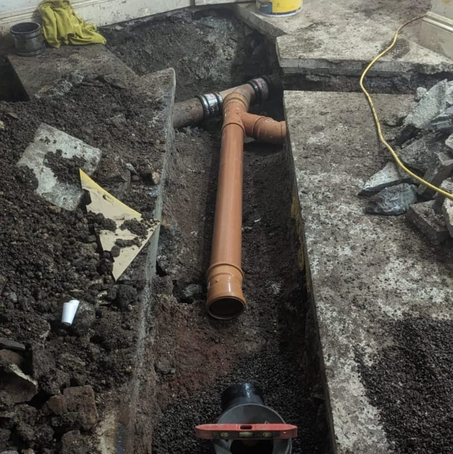 A construction site with exposed underground plumbing, featuring a newly installed brown PVC pipe in a T-junction configuration. Surrounding the pipe is dug-up dirt and gravel. Various construction tools and materials are scattered nearby, including a saw and a yellow cord.