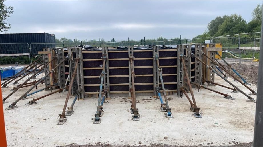 A construction site featuring a large rectangular concrete formwork structure surrounded by metal bracing supports and scaffolding. The setup is on a flat concrete base with a cloudy sky and trees in the background.