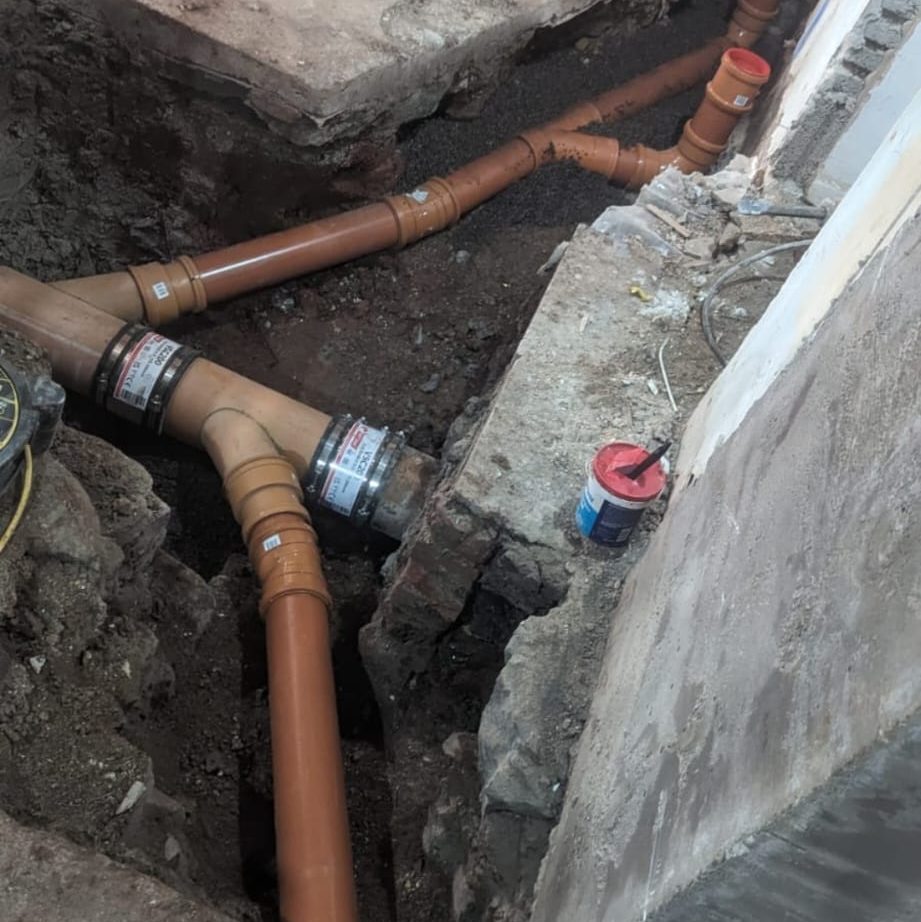 A construction site showing installed drainage pipes with brown fittings and a gray rocky surrounding. There is a red-and-black container and some wires on the right side near a partially built wall.