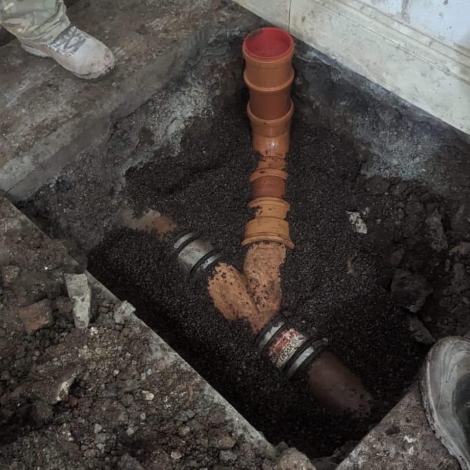 A construction site showing an open ground excavation with a newly installed pipe junction. The pipes are surrounded by gravel and soil, and workers' feet are visible near the edge of the pit. The pipes are connected with couplers and lead in various directions.