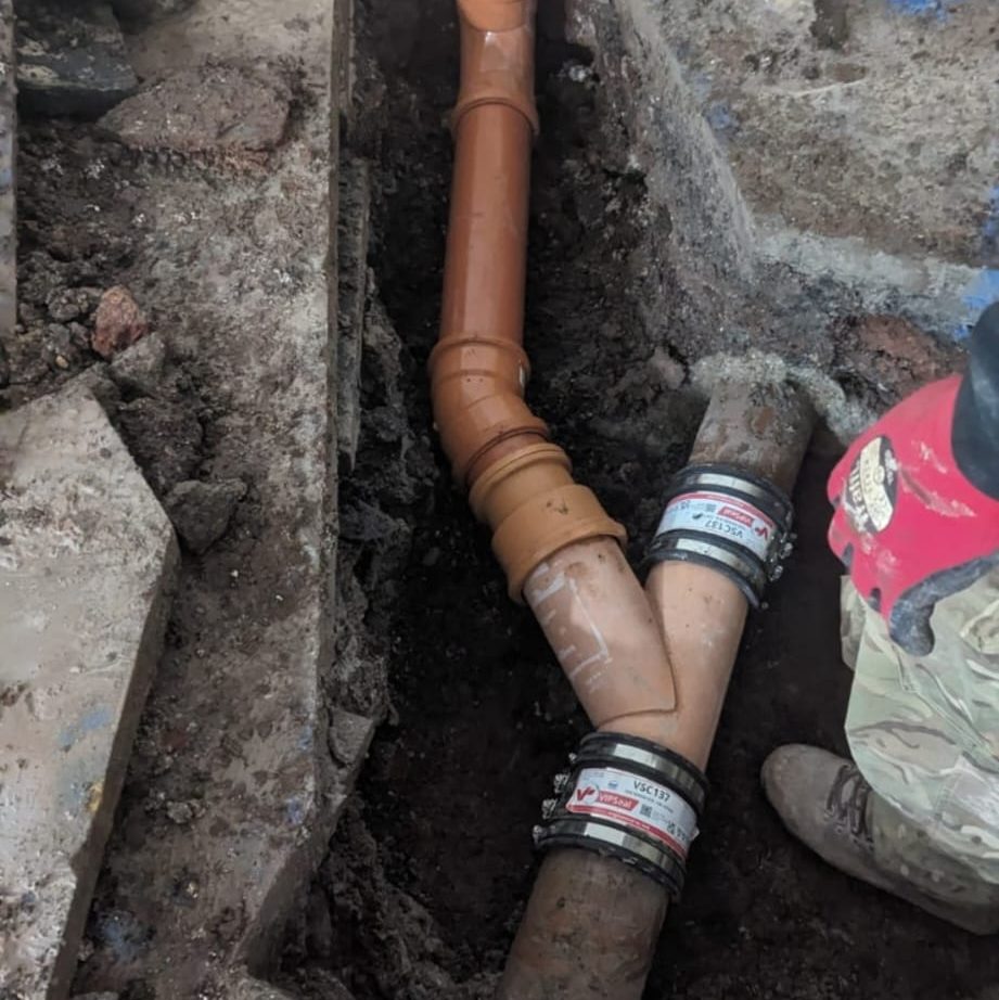 A technician with red gloves is working on a repaired sewer line in a dug-up area. The pipes are connected with coupling sleeves, and the ground is visibly disturbed from the excavation work. Only the lower half of the technician's body is visible.