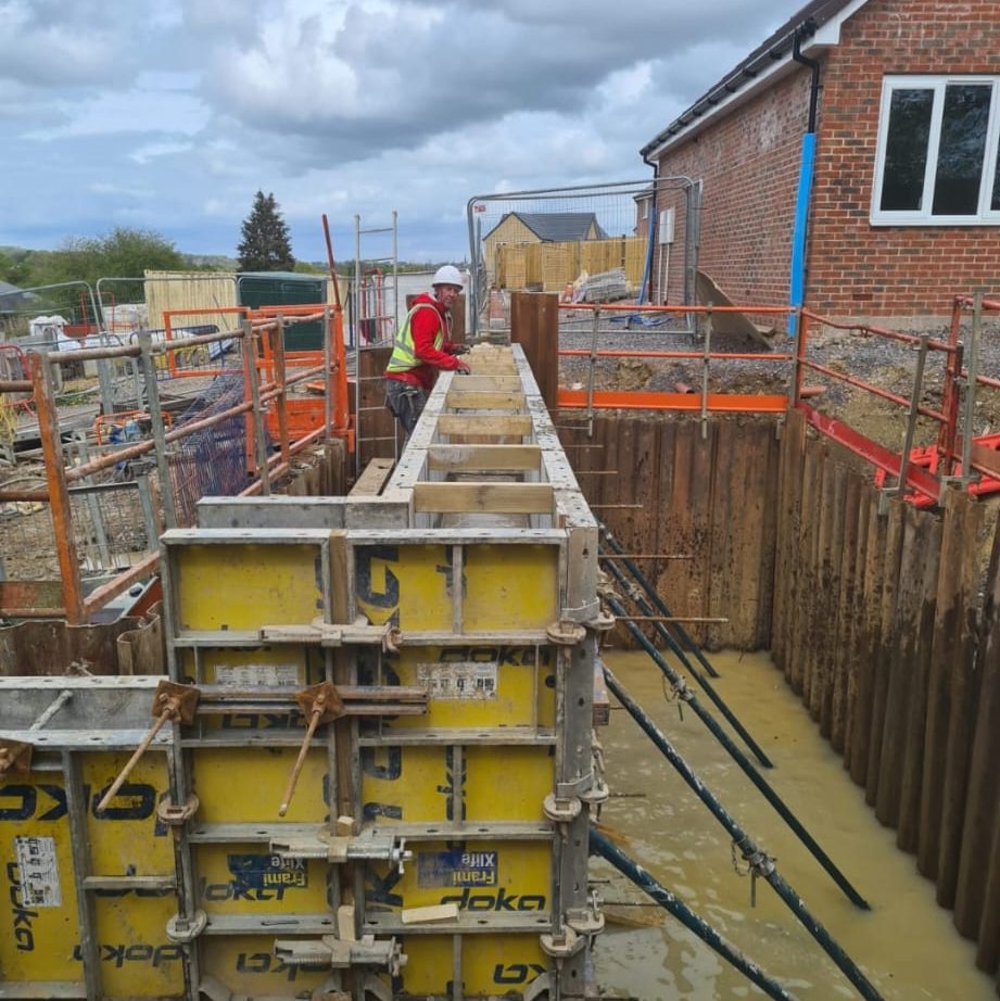 A construction worker in a red jacket and yellow safety vest is aligning foundation wall forms at a muddy construction site. The building behind is a red-brick structure with white-framed windows. Various construction materials and safety barriers surround the area.