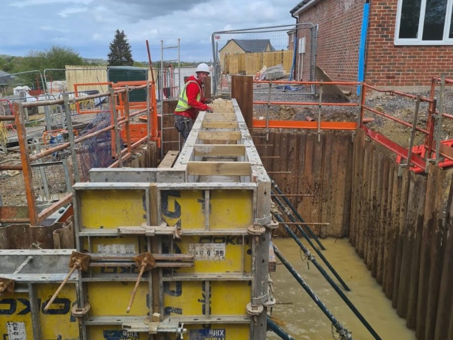 Construction worker on a site overlooking a partially completed structure with metal formwork in place for a concrete foundation. Worker is wearing a red and yellow safety vest and white hard hat. The site is fenced off and near a brick building.