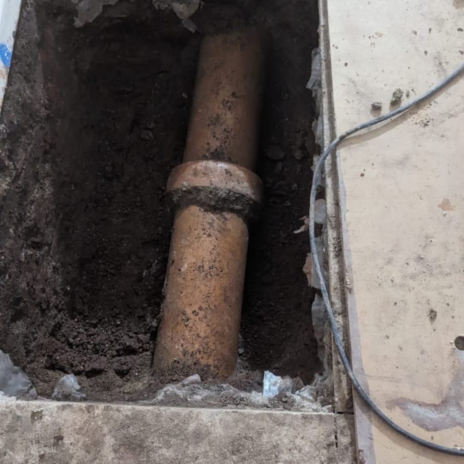 A partially exposed clay pipe runs horizontally through a dug-out section in a concrete floor. The area surrounding the pipe is filled with soil, and electrical wiring extends along the right side of the image.