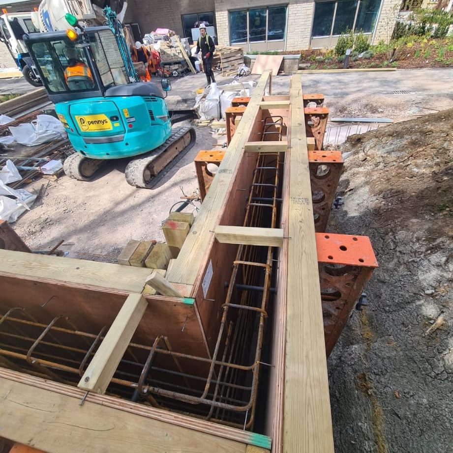 A construction site with a blue excavator in the background. In the foreground, wooden and metal formwork structures are being prepared for pouring concrete. Workers in safety gear are visible, and a building under construction is seen in the background.