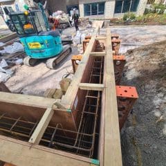 Construction site with a concrete form frame for a structure in the foreground. A small blue excavator marked with "JCB" is parked nearby. Workers and building materials are scattered around, and a building under construction is visible in the background.