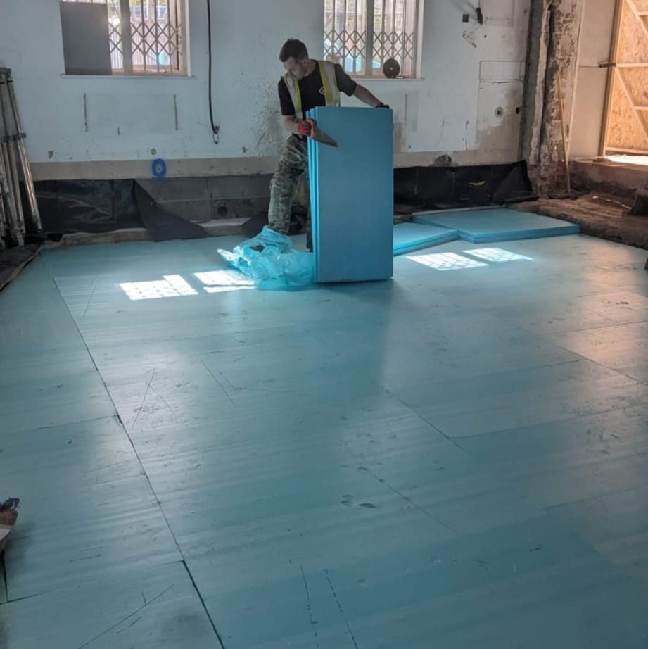 A construction worker in a safety vest is installing or positioning blue insulation panels on a floor in a room under renovation. The room has exposed walls and windows with security bars. The floor is partially covered with the blue panels.