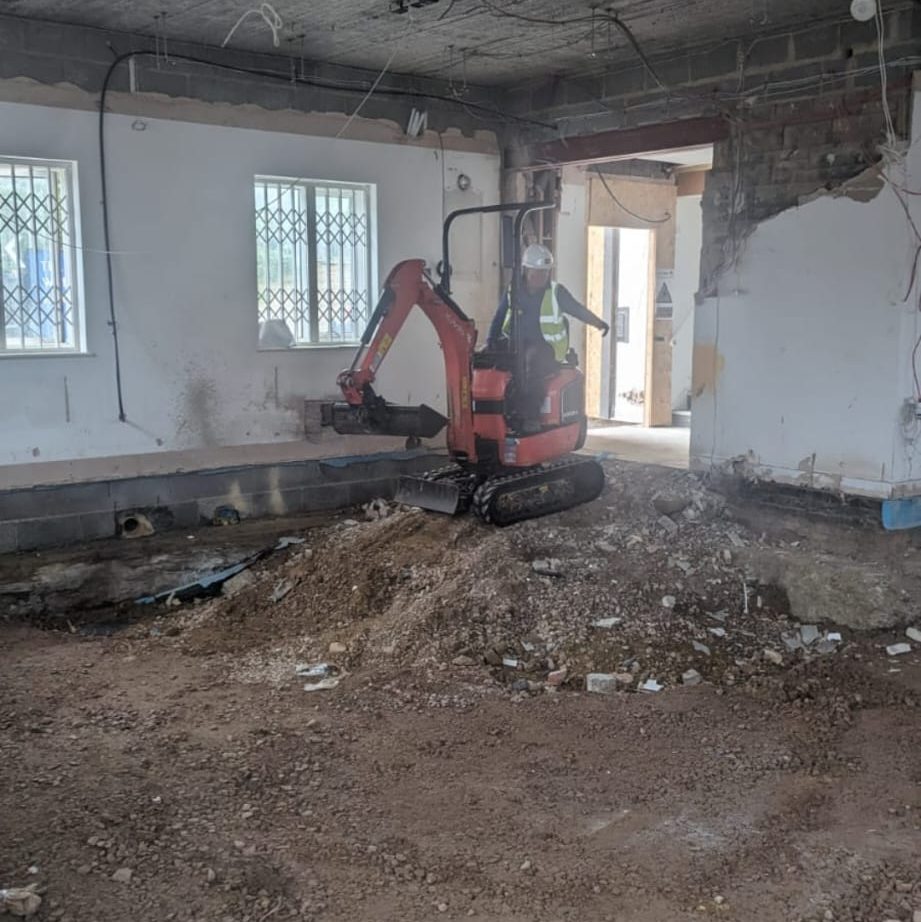 A small excavator operates inside a partially demolished room, handling a pile of rubble and dirt. Walls are stripped to the studs in some areas, and exposed wiring is visible. A worker in protective gear is driving the excavator. Light enters through barred windows.