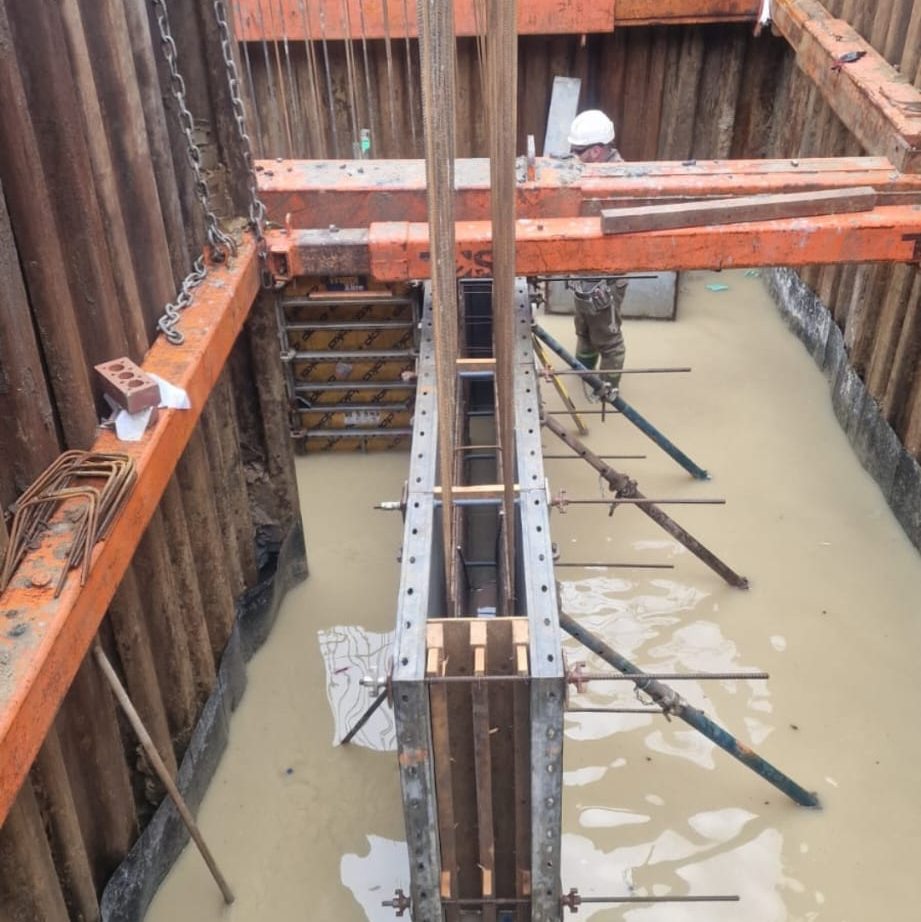 A construction site showing a deep, rectangular excavated pit partially filled with water. Vertical metal scaffold structures are in place, and a worker wearing a white hard hat and safety gear is present, with wooden support beams and metal rods around the area.