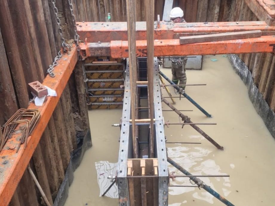 A construction worker in a white helmet stands in a flooded construction site, surrounded by wooden and metal supports. The worker appears to be aligning or inspecting a vertical metal framework positioned at the center of the site.