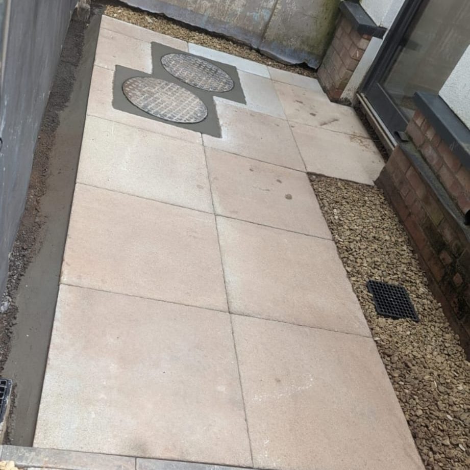 An outdoor walkway with large, square, beige tiles. Two circular manhole covers are integrated into the tiles near the center. The path is flanked by walls and edges lined with small gravel. A sliding door is partially visible on the right.