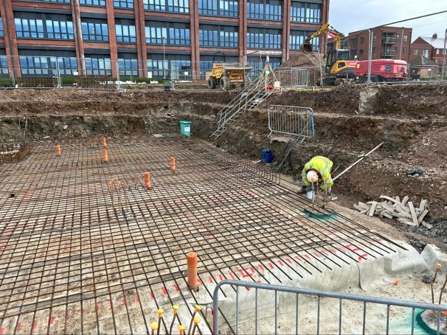 Construction site featuring a large excavation area with steel rebar grid laid out across the ground. A worker in high-visibility clothing is positioning steel rods. Various construction vehicles and equipment are in the background, with a partially built structure nearby.