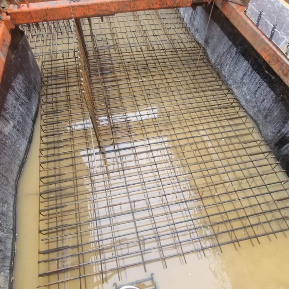 Reinforcement steel bars arranged in a grid pattern are submerged in muddy water within a construction site. The partially completed concrete formwork is visible around the bars, indicating ongoing foundation work.