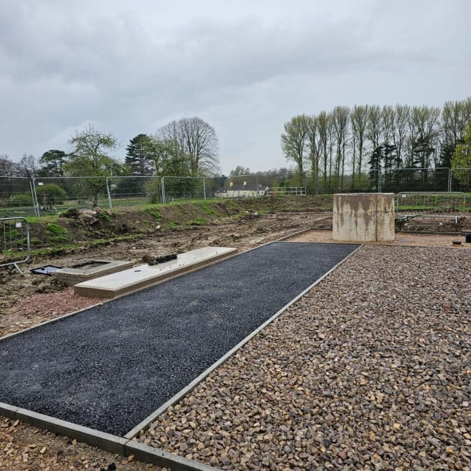 A construction site with a large gravel and asphalt area, surrounded by trees, fencing, and some equipment. In the background, more trees and a cloudy sky are visible. There is also a concrete slab and a small concrete structure on the ground.