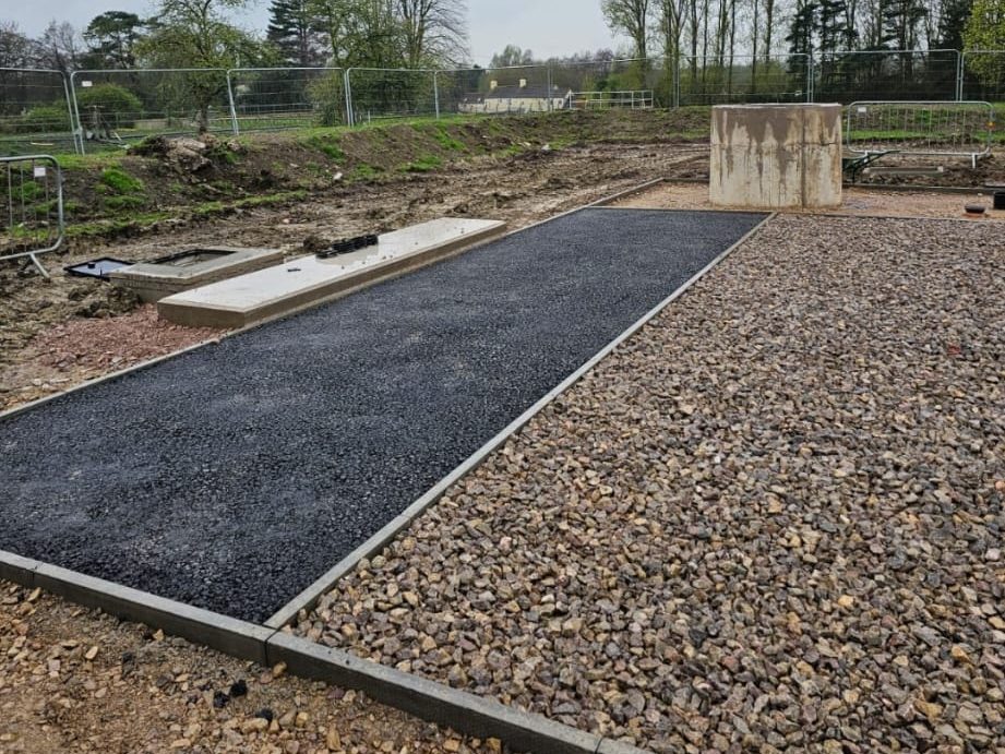 A construction site with a freshly laid asphalt path bordered by gravel. Metal fencing surrounds the area. Trees and a building are visible in the background. Construction materials are scattered around, and the ground appears uneven and muddy.