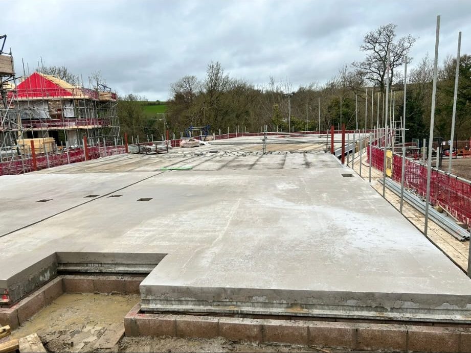 A construction site with a large concrete foundation slab, scaffolding on the sides, and some wooden structures in the background. The area is surrounded by trees and the sky is cloudy.