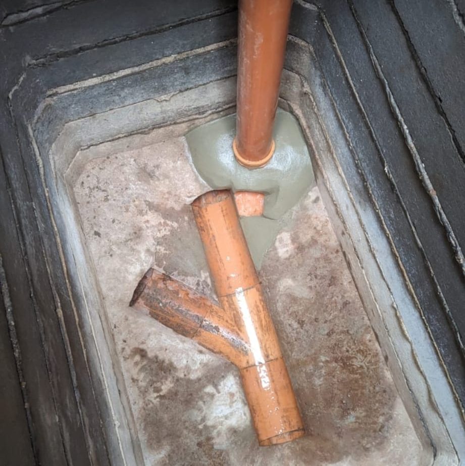 An underground plumbing installation featuring a Y-shaped brown pipe connected to an upright orange pipe. The pipes are set in a cemented rectangular pit with concrete walls. The pipes appear to be part of a drainage or sewage system.