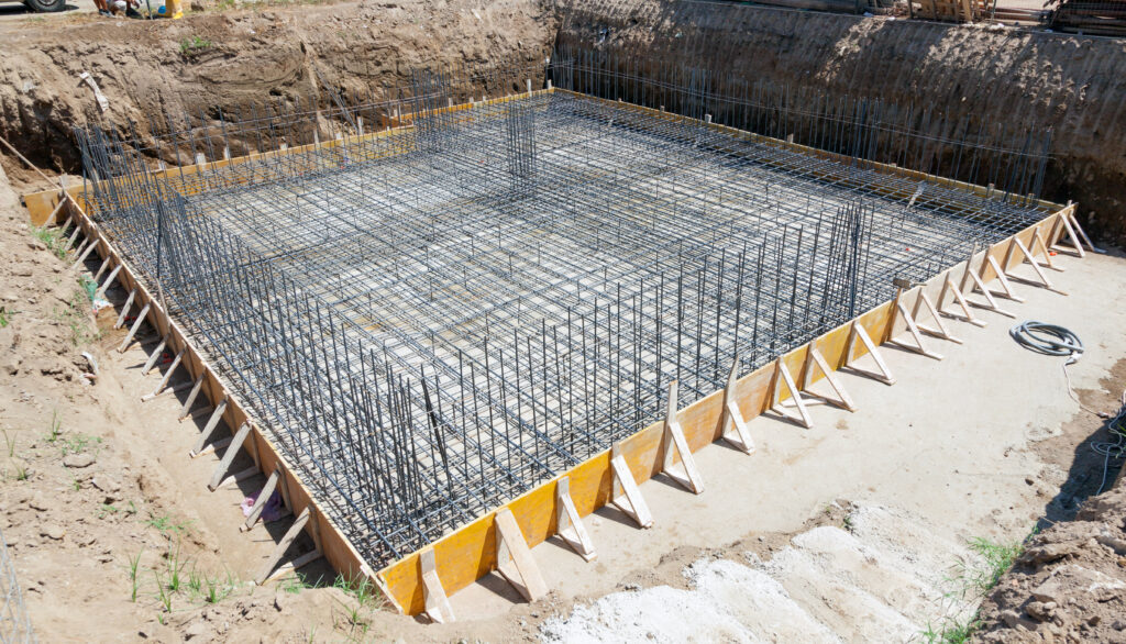 A construction site showing a square foundation with steel reinforcement bars and wooden formwork, prepared for pouring concrete in an excavated area surrounded by soil.