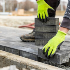 A person wearing bright green gloves is placing rectangular paving stones to create a walkway outdoors. The worker is on a construction site with other materials and workers blurred in the background.
