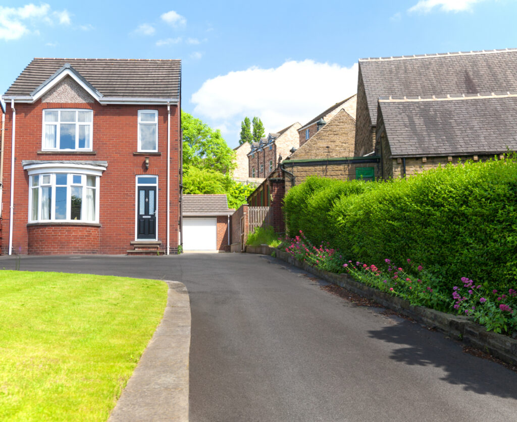 A brick house with white trim and a bay window stands next to a paved driveway bordered by green hedges and pink flowers, with other buildings in the background under a blue sky.