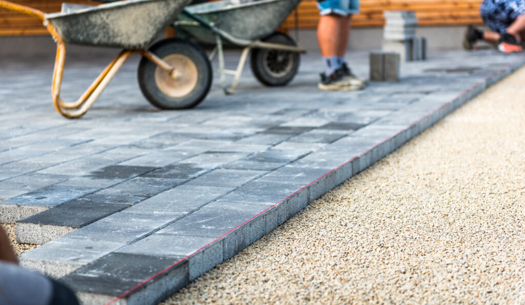 Workers are laying gray concrete paving stones on a construction site. A wheelbarrow is nearby, and a string line marks the alignment. Gravel is seen in the foreground, with some paving stones stacked in the background.
