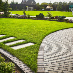 Curved brick paths wind through a well-maintained green lawn with ornamental plants, stone steps, and small soccer goals in the background, suggesting a landscaped garden or park setting.