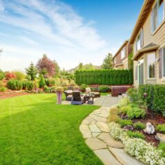 A spacious backyard with a manicured lawn, stone path, patio furniture, and landscaped garden beds beside a tan house under a clear blue sky. Shrubs, flowers, and trees add vibrant color to the scene.