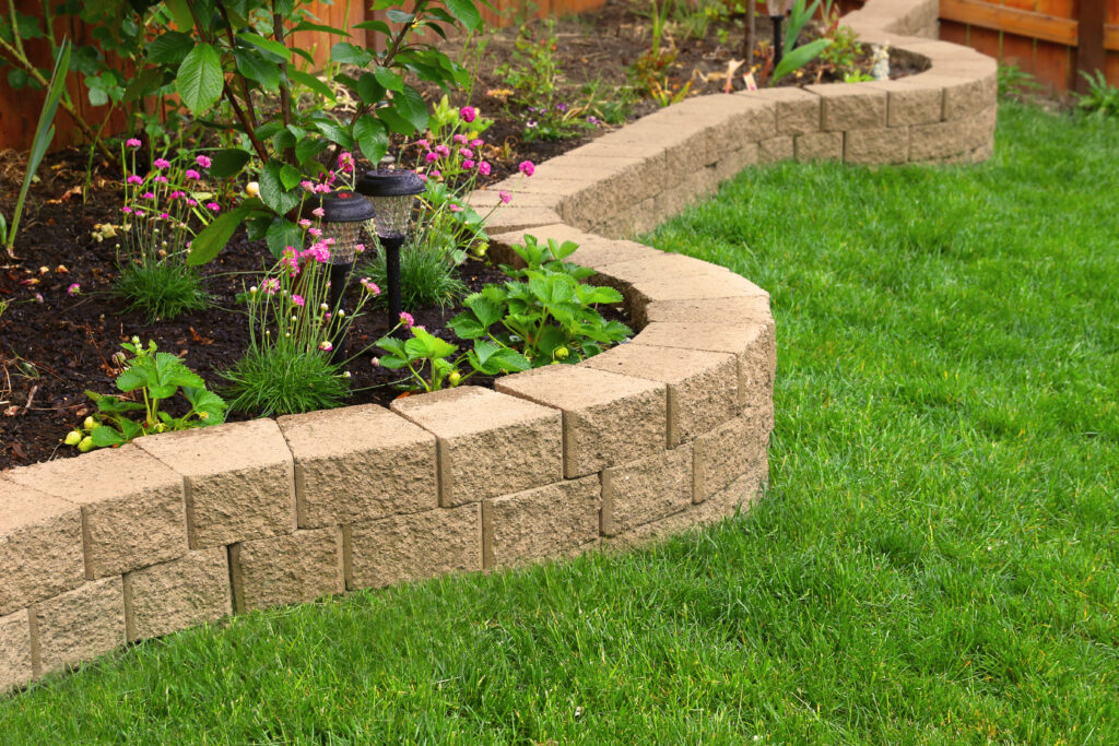 A curved stone retaining wall separates a garden bed with flowers and plants from a neatly trimmed green lawn in a backyard.