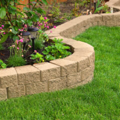 A curved stone retaining wall separates a garden bed with flowers and plants from a neatly trimmed green lawn in a backyard.