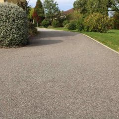 A smooth, freshly laid asphalt driveway curves through a well-maintained garden with green grass, bushes, and trees on a sunny day.