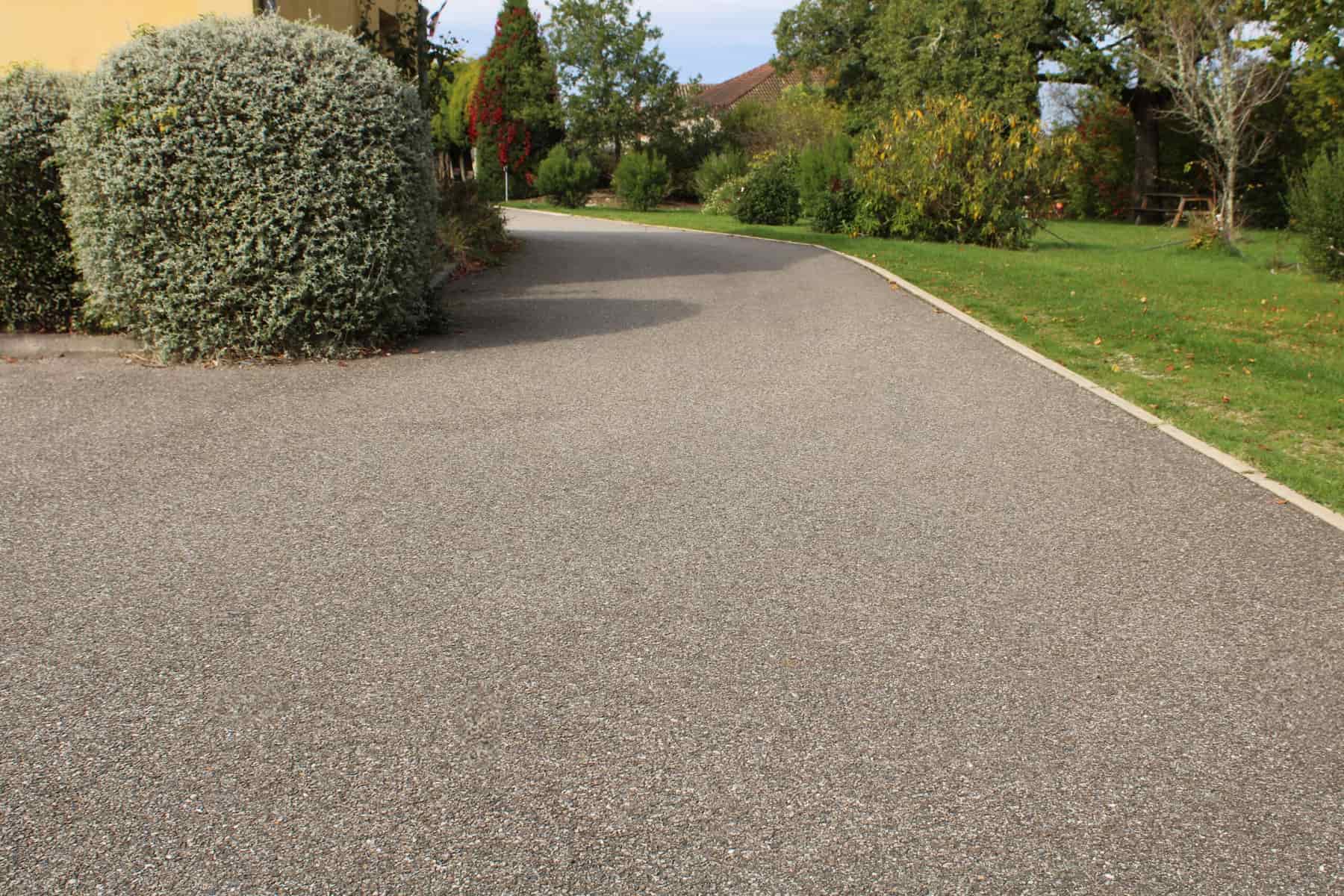 A smooth, freshly laid asphalt driveway curves through a well-maintained garden with green grass, bushes, and trees on a sunny day.