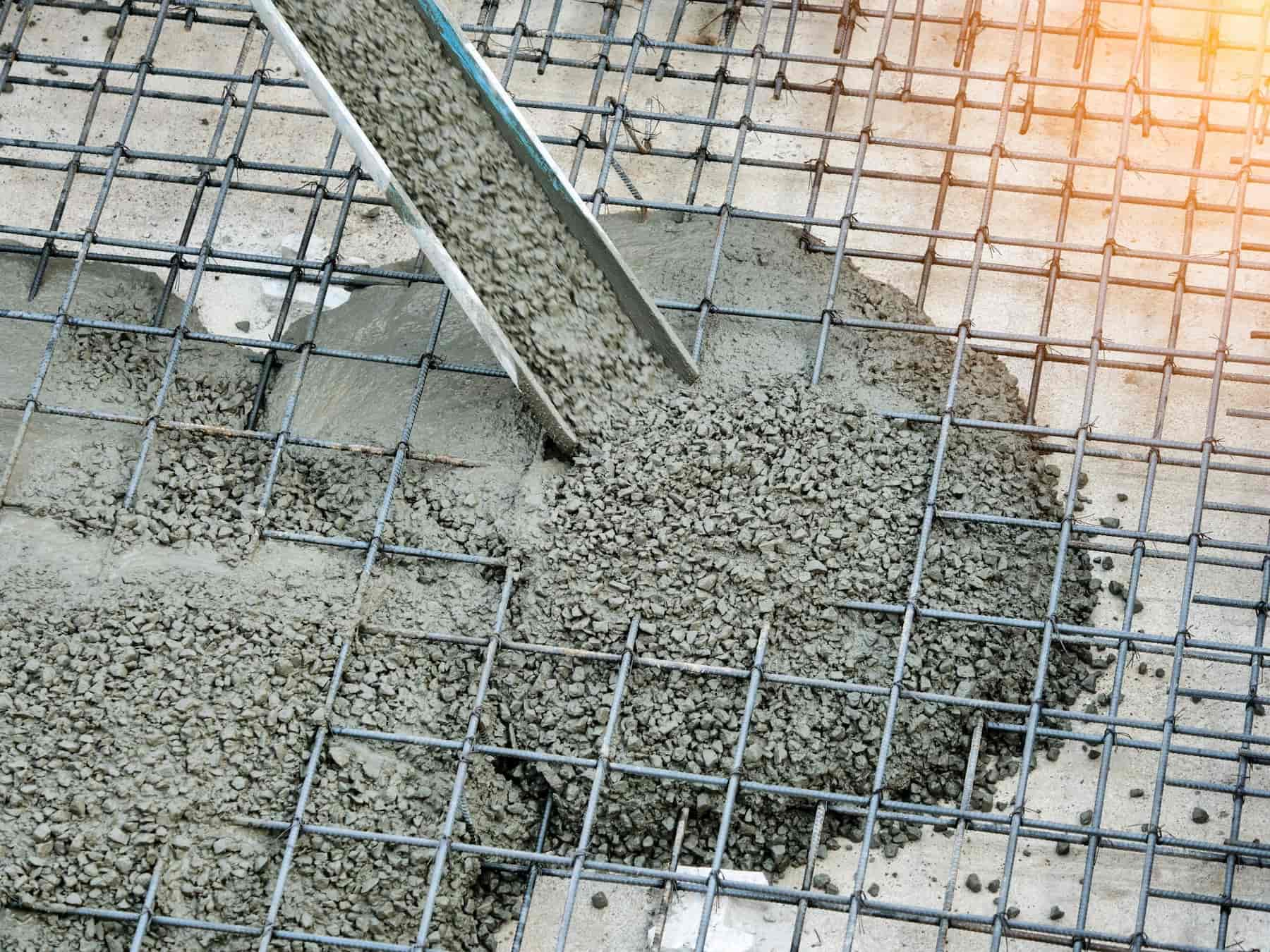 Wet concrete being poured onto a steel rebar reinforcement grid, part of a construction site foundation, with sunlight shining from the top right corner.