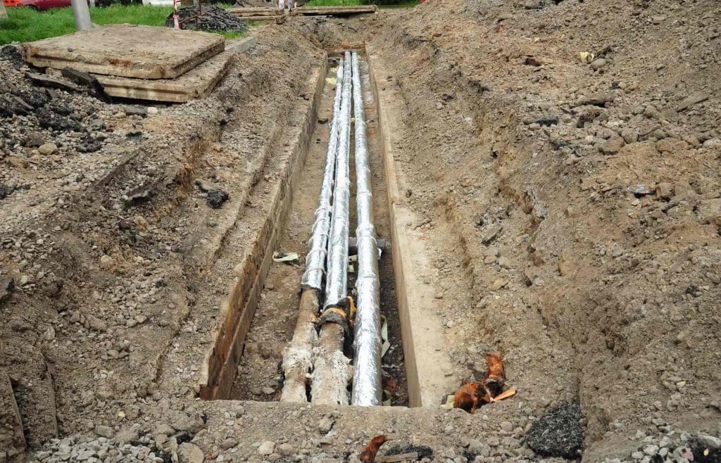 A trench dug in the ground contains three insulated pipes running parallel, partially covered with soil, surrounded by dirt and construction materials at a work site.